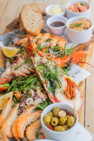Platter of seafood with a focus on a lobster, surrounded by bread, sauces, and olives on a wooden board.